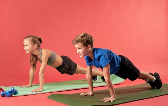 Cute girl and boy in sportswear holding push-up position and smiling while doing strength exercise on yoga mats