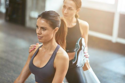 Exercising with fitness instructor. Young beautiful caucasian woman doing stretching exercises with assistance of her personal trainer at gym. Sport and healthy lifestyle concept