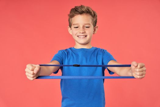 Adorable male child holding resistance exercise band and smiling while standing against red background