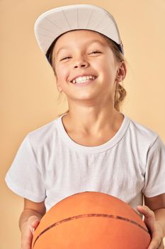 Adorable girl basketball player looking at camera and smiling while standing against light orange background