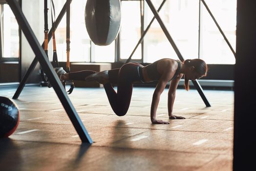 Suspension Training. Side view of young caucasian fitness woman in sportswear doing TRX workout at industrial gym, working out with fitness straps. Sport, wellness and healthy lifestyle