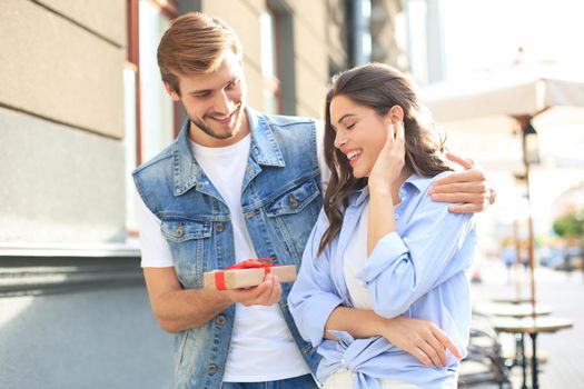 Astonished excited couple in summer clothes smiling and holding present box together while standing on city street