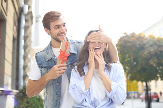 Astonished excited couple in summer clothes smiling and holding present box together while standing on city street