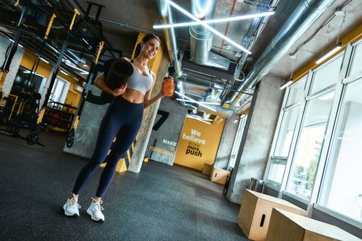 Full length of a young beautiful fitness woman in sportswear holding yoga mat and bottle of water, looking at window and smiling while exercising at gym. Sport, wellness and healthy lifestyle