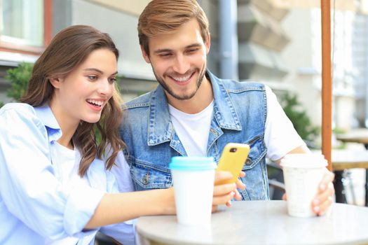 Attractive young couple in love drinking coffee while sitting at the cafe table outdoors, using mobile phone