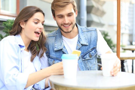 Attractive young couple in love drinking coffee while sitting at the cafe table outdoors, using mobile phone