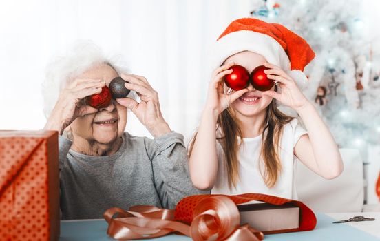 Smiling granddaughter in santa hat holding decorative balls like eyes with grandma at Christmas