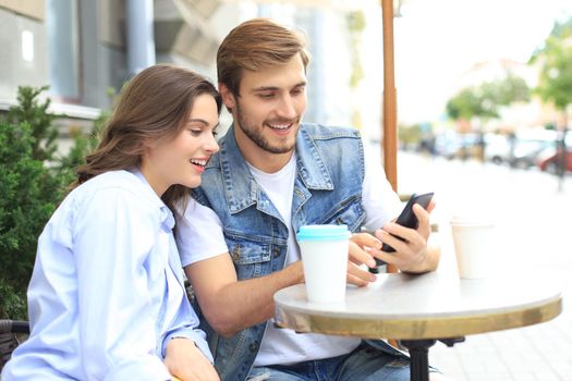 Attractive young couple in love drinking coffee while sitting at the cafe table outdoors, using mobile phone