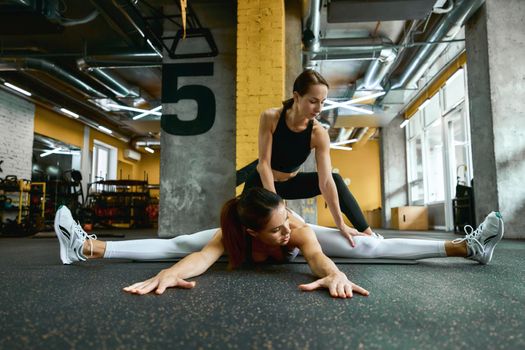 Young beautiful fitness woman in sportswear sitting on yoga mat at gym and doing stretching exercises with assistance of her personal trainer. Sport and healthy lifestyle concept