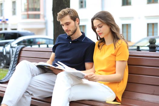 Romantic young couple in summer clothes smiling and reading books together while sitting on bench in city street