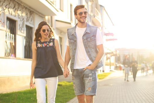 Image of lovely happy couple in summer clothes smiling and holding hands together while walking through city street