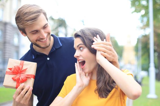 Astonished excited couple in summer clothes smiling and holding present box together while standing on city street