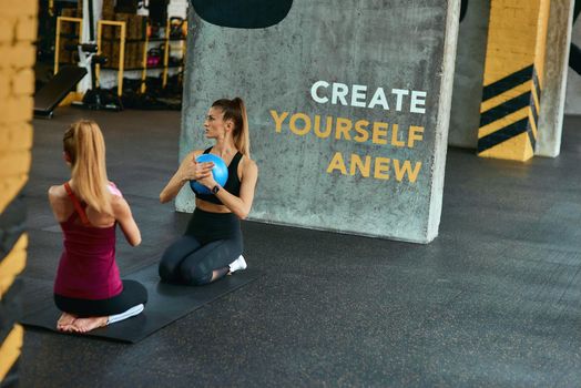 Pilates class. Two young athletic women in sportswear exercising with small fitness ball while sitting on yoga mat on the floor at gym. Sport, training, wellness and healthy lifestyle