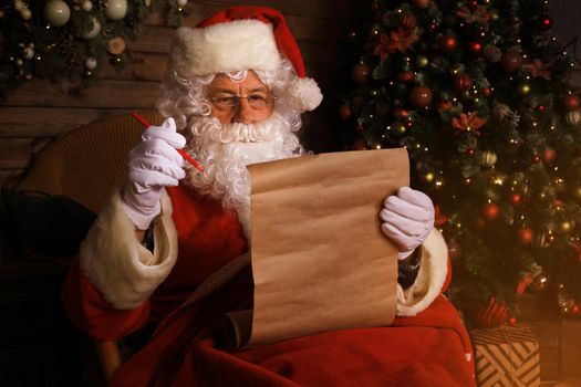 Portrait of happy Santa Claus sitting at his room at home near Christmas tree and reading Christmas letter or wish list