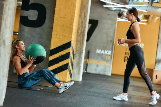 Young athletic woman in sportswear exercising with fitness ball at gym with assistance of personal trainer, doing abs exercises. Sport, training, wellness and healthy lifestyle