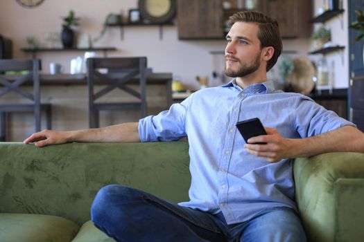 Attractive young man relaxing on a couch at home and using mobile phone for cheking social nets