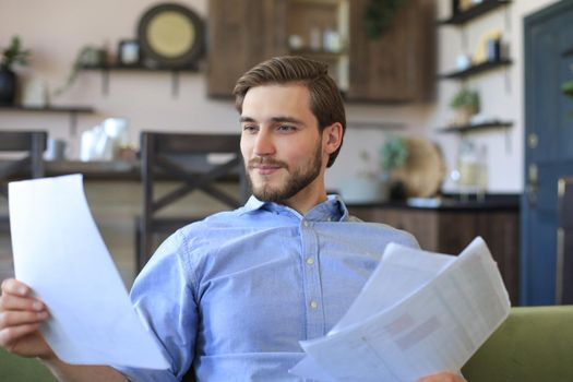 Concentrated young freelancer businessman sitting on sofa with laptop and examining documents