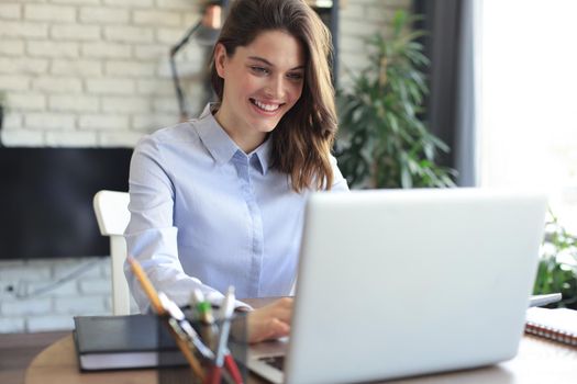 Smiling pretty woman sitting at table, looking at laptop screen. Happy entrepreneur reading message email with good news, chatting with clients online