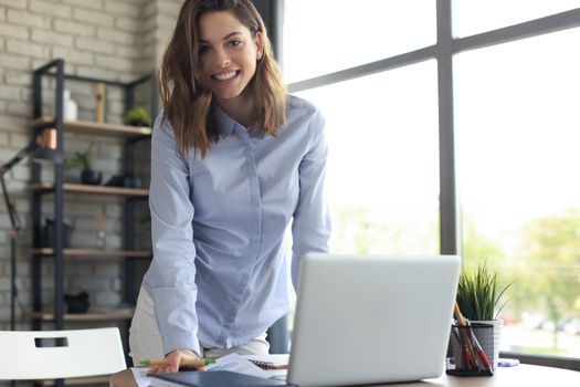 Confident young businesswoman with a friendly smile standing behind her desk in a home office looking at the camera
