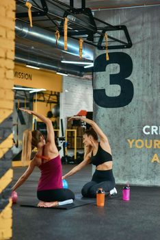 Stretching exercises. Vertical shot of two young athletic women in sportswear warming up before workout while sitting on the floor at gym. Sport, training, wellness and healthy lifestyle