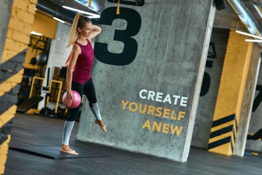 Full length of a young caucasian woman in sportswear exercising with small fitness ball at modern gym. Sport, workout, wellness and healthy lifestyle