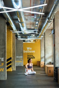 Vertical shot of a young athletic woman in sportswear sitting on yoga mat at gym and looking in window, warming up or stretching body before workout. Sport, wellness and healthy lifestyle