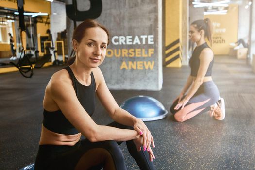 Young beautiful woman in sportswear smiling at camera while working out with friend at gym, using special fitness equipment. Healthy lifestyle, training, wellness and bodycare