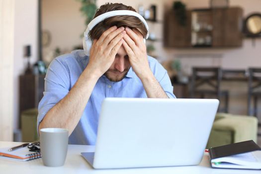 Unhappy frustrated young male holding head by hands sitting with laptop behind desk at home