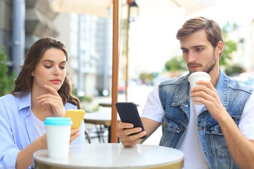 Attractive young couple in love drinking coffee while sitting at the cafe table outdoors, using mobile phone