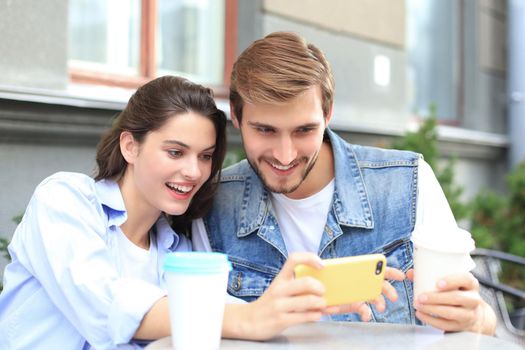 Attractive young couple in love drinking coffee while sitting at the cafe table outdoors, using mobile phone