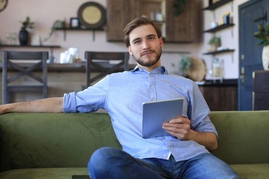 Smiling handsome man holding and using tablet with social networks at home, happy freelancer making online shopping, sitting on cozy sofa at living room