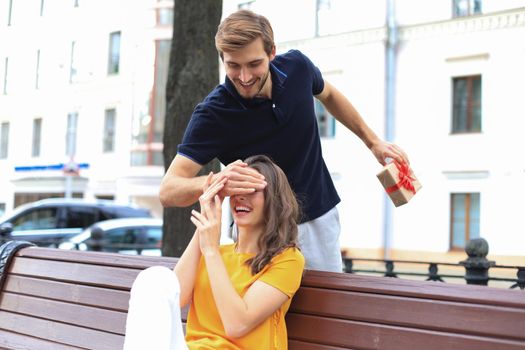 Image of charming excited couple in summer clothes smiling and holding present box together while sitting on bench