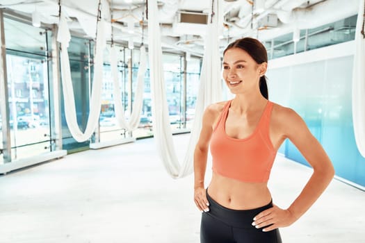 Healthy living. Young happy woman in sportswear, female fitness instructor keeping arms on hips and smiling while standing in a beautiful fly yoga studio. Sport, wellness, active lifestyle