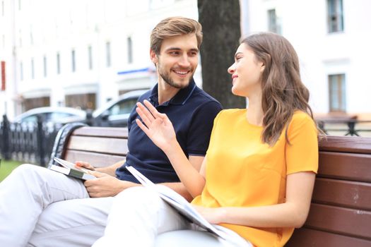 Romantic young couple in summer clothes smiling and reading books together while sitting on bench in city street