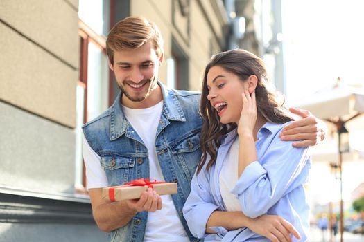 Astonished excited couple in summer clothes smiling and holding present box together while standing on city street