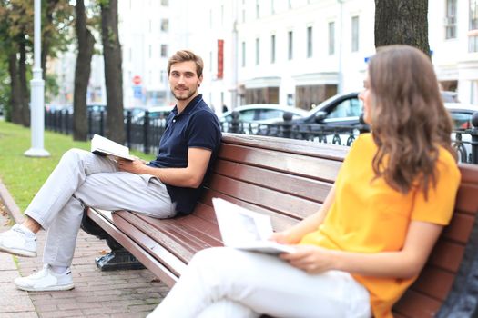 Romantic young couple in summer clothes smiling and reading books together while sitting on bench in city street