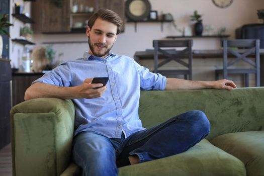 Attractive young man relaxing on a couch at home and using mobile phone for cheking social nets