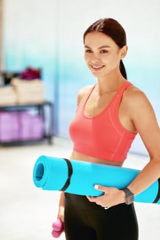 Young attractive woman in sportswear holding yoga mat and smiling while standing in studio before yoga class or fitness training, vertical shot. Sport, wellness and healthy lifestyle