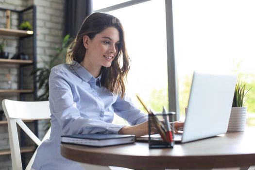 Smiling pretty woman sitting at table, looking at laptop screen. Happy entrepreneur reading message email with good news, chatting with clients online
