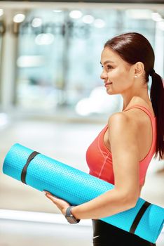 Side view of a young attractive woman in sportswear holding yoga mat and looking away while standing in studio before yoga class or fitness training. Sport, wellness and healthy lifestyle