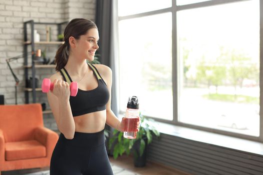 Young beautiful woman standing with dumbbell and drink water after exercising. Attractive female bodybuilder working out. Fitness and healthy lifestyle concept.