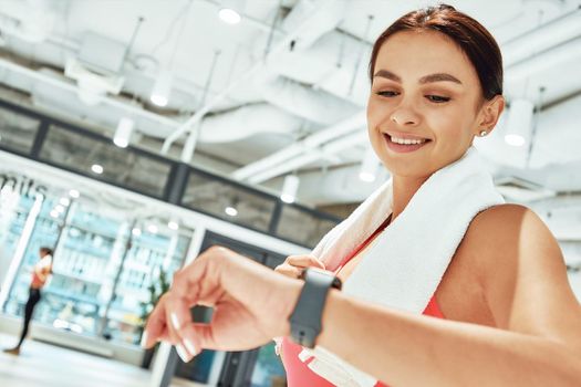 Young happy sportive woman with white towel on shoulders looking at smartwatch while exercising in fitness studio. Sport, workout, wellness and healthy lifestyle concept