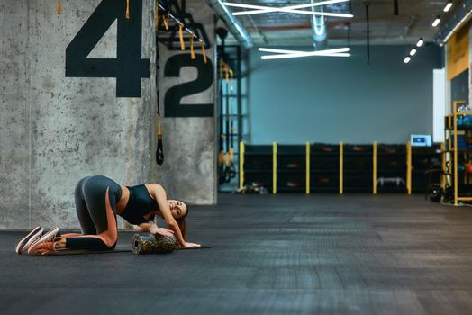 Full length of a young sportive woman in sportswear exercising with foam roller, doing stretching exercises on yoga mat at industrial gym. Sport, training, workout and healthy lifestyle