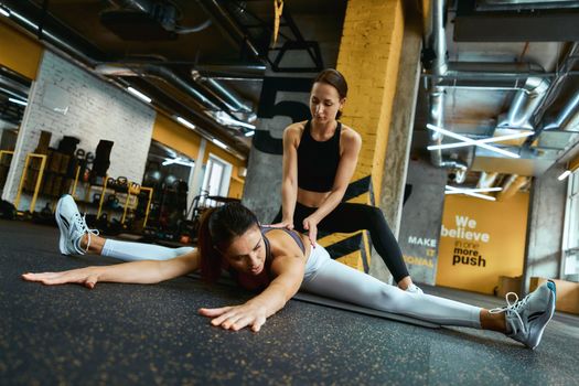 Stretching exercises. Young flexible fitness woman in sportswear practicing transverse twine with assistance of her personal trainer at gym. Sport and healthy lifestyle concept