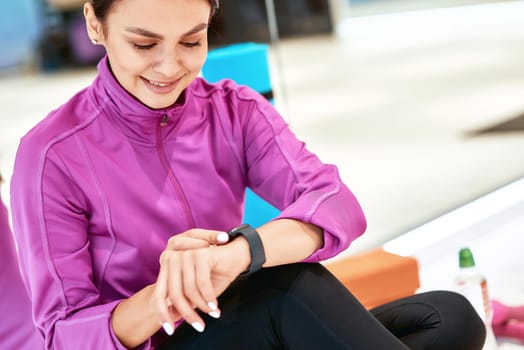 Close up shot of a young beautiful woman in sportswear sitting on the floor in studio or gym and looking at smartwatch. Resting after fitness training. Sport, wellness and healthy lifestyle