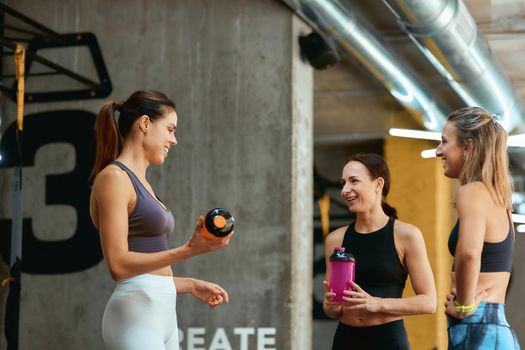 Resting after workout. Group of three young beautiful fitness women in sportswear talking and smiling while taking a break at gym. Sport, wellness and healthy lifestyle