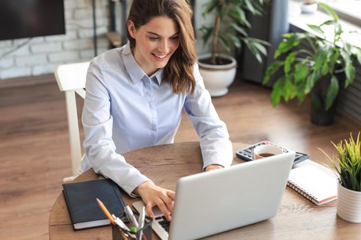 Smiling pretty woman sitting at table, looking at laptop screen. Happy entrepreneur reading message email with good news, chatting with clients online