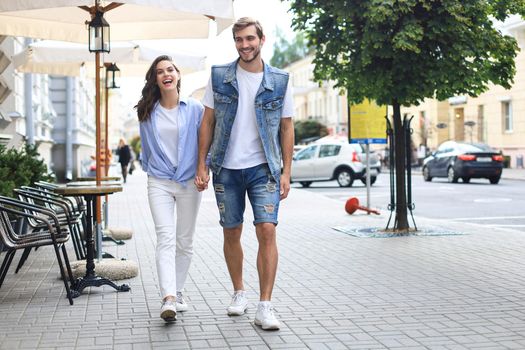 Image of lovely happy couple in summer clothes smiling and holding hands together while walking through city street