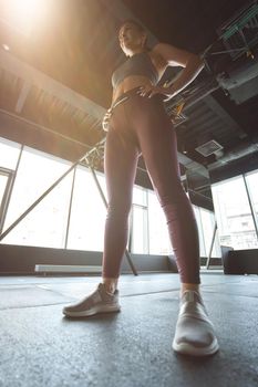 Bottom view of a strong athletic woman in sportswear keeping arms on hips while working out at industrial gym, vertical shot. Sport, wellness and healthy lifestyle