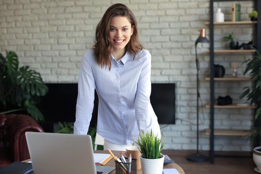 Confident young businesswoman with a friendly smile standing behind her desk in a home office looking at the camera